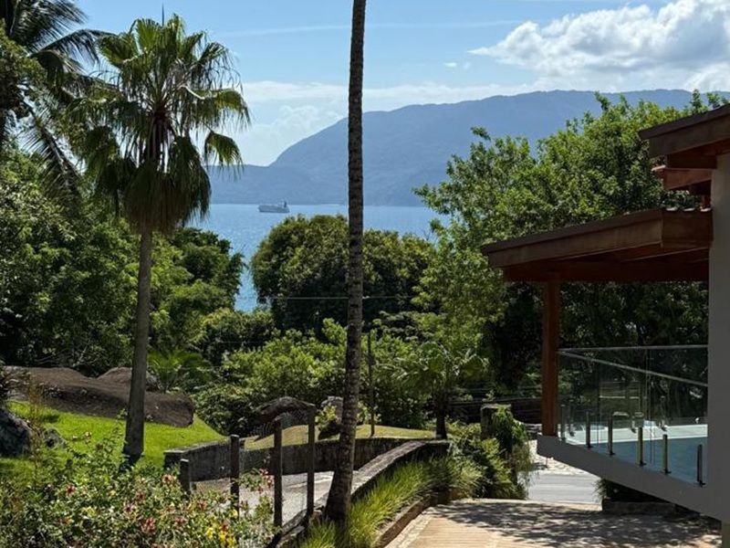 Uma vista panorâmica com vista para árvores verdejantes, um mar calmo e montanhas ao longe, com uma varanda moderna em primeiro plano.