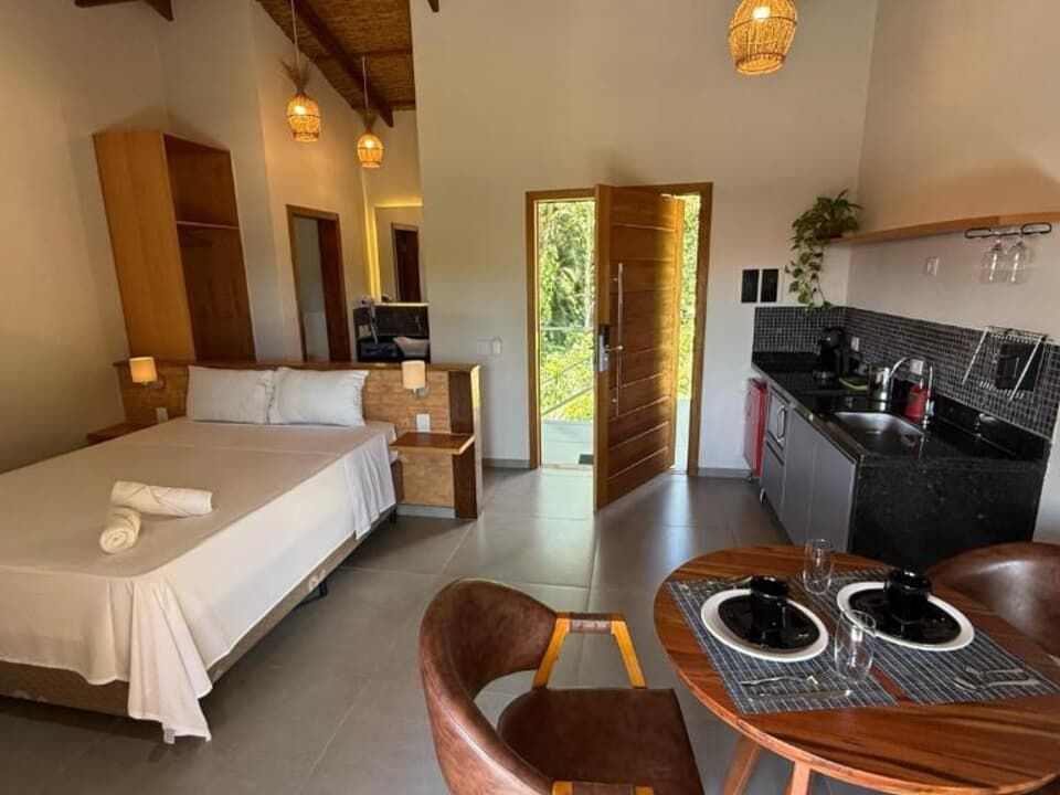 Um quarto de hotel com cama feita de branco, uma pequena cozinha com bancadas pretas e um conjunto de jantar de madeira.