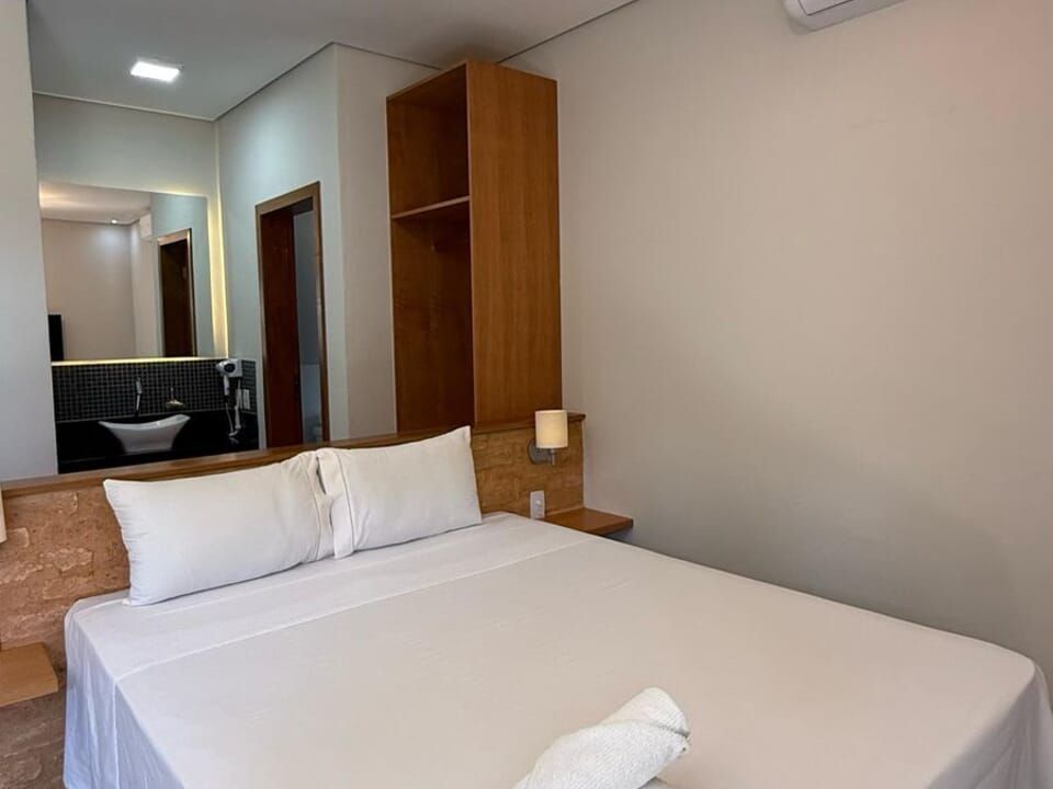 Um quarto de hotel com cama queen-size, lençóis brancos, prateleiras em tons de madeira, um abajur e vista para um banheiro com espelho.