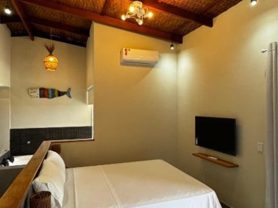 Um quarto com uma cama de lençóis brancos, uma TV fixada na parede, um peixe decorativo na parede e um teto inclinado de palha.