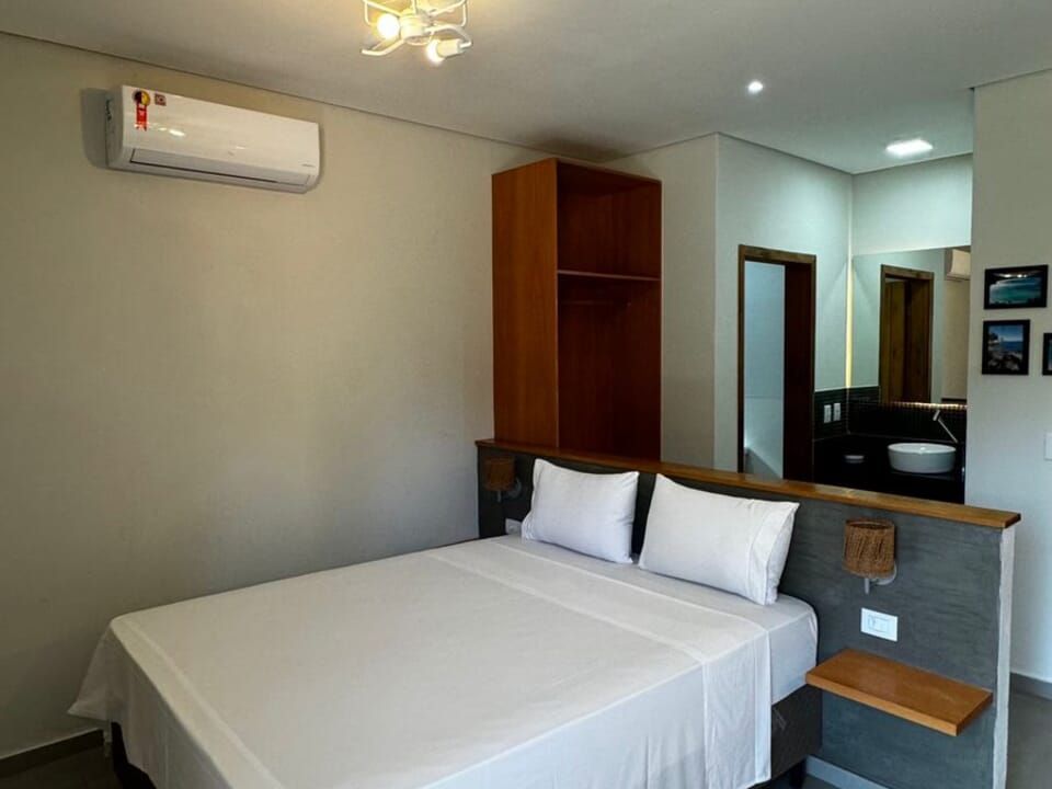 Um quarto de hotel com cama de lençóis brancos, guarda-roupa de madeira, ar-condicionado de parede e banheiro aberto.