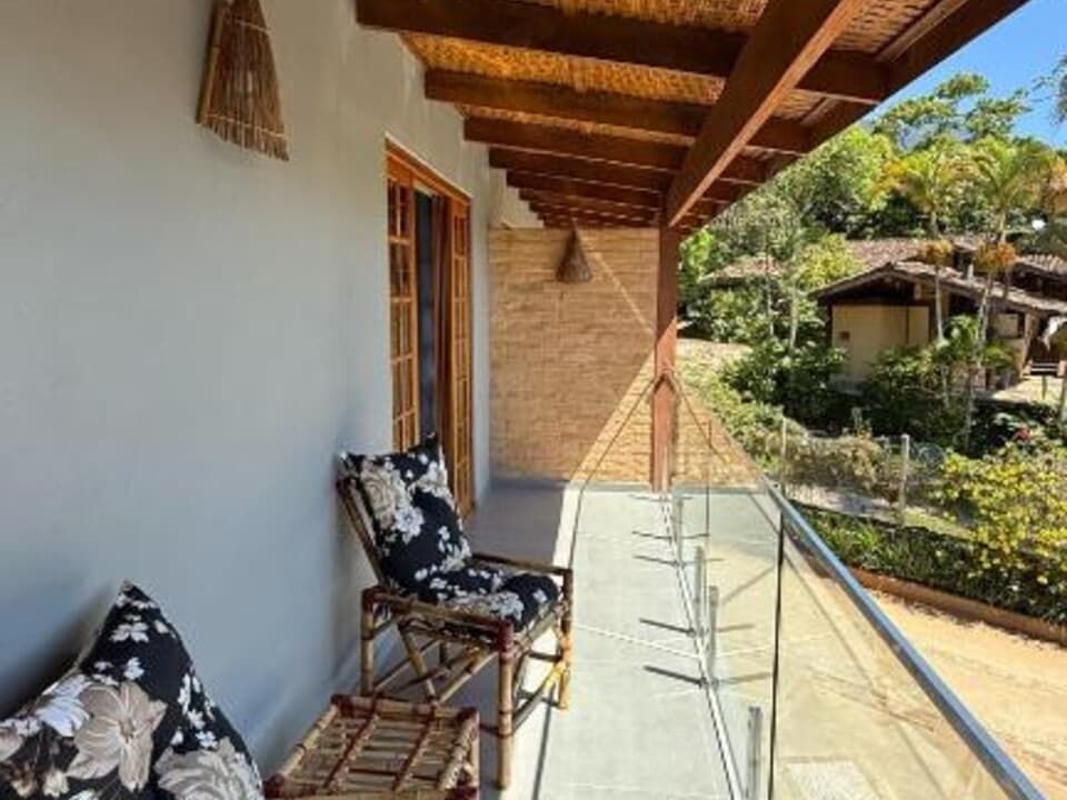 Uma varanda com teto de madeira, guarda-corpo de vidro e poltronas com estampa floral, com vista para uma paisagem tropical.
