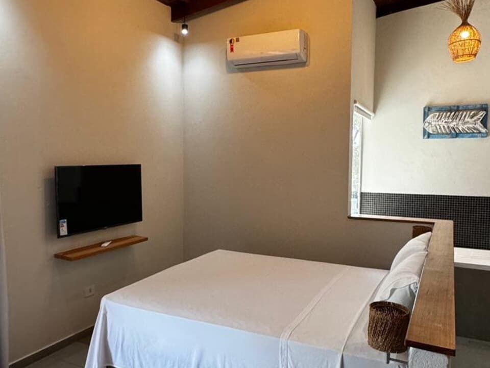 Interior de um quarto de hotel com cama de lençóis brancos, TV de parede, ar-condicionado split e decoração com temática praiana.