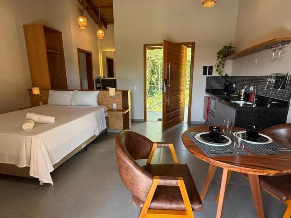 Um apartamento estúdio com cama de casal com lençóis brancos, uma pequena mesa de jantar com cadeiras e um balcão de cozinha.