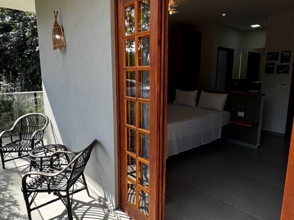 Uma varanda com duas cadeiras de vime pretas e uma pequena mesa dá para um quarto com uma cama e móveis escuros.