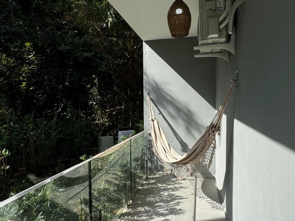 Uma rede bege está pendurada em uma varanda com parapeito de vidro, com vista para uma exuberante área florestal verde em um dia ensolarado.