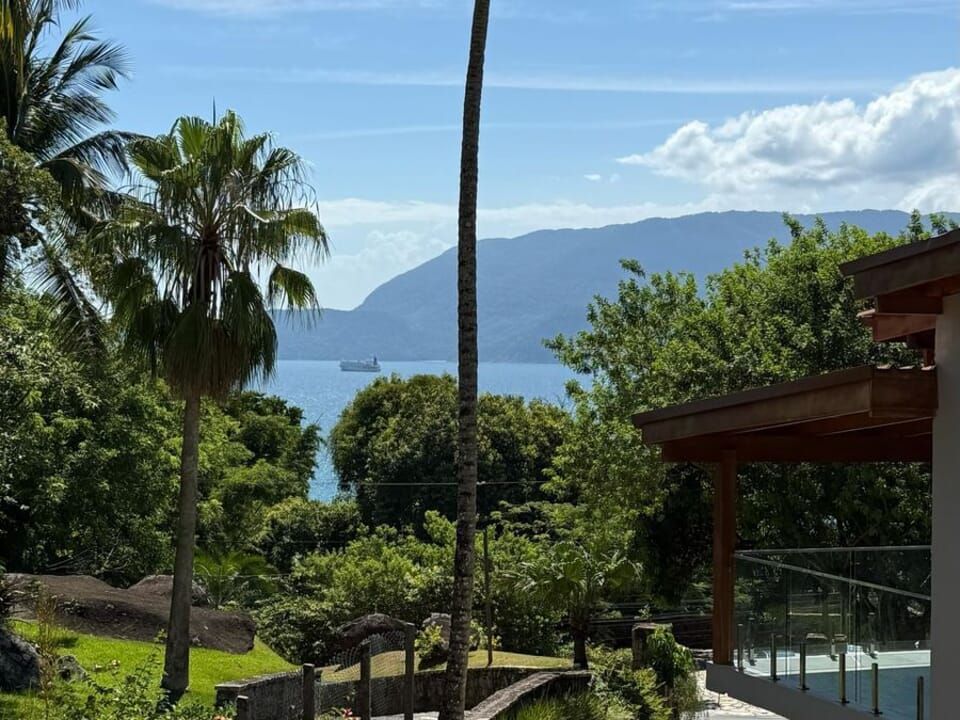 Uma paisagem exuberante com palmeiras e folhagem verdejante, com vista para uma baía e montanhas ao longe, sob um céu azul.