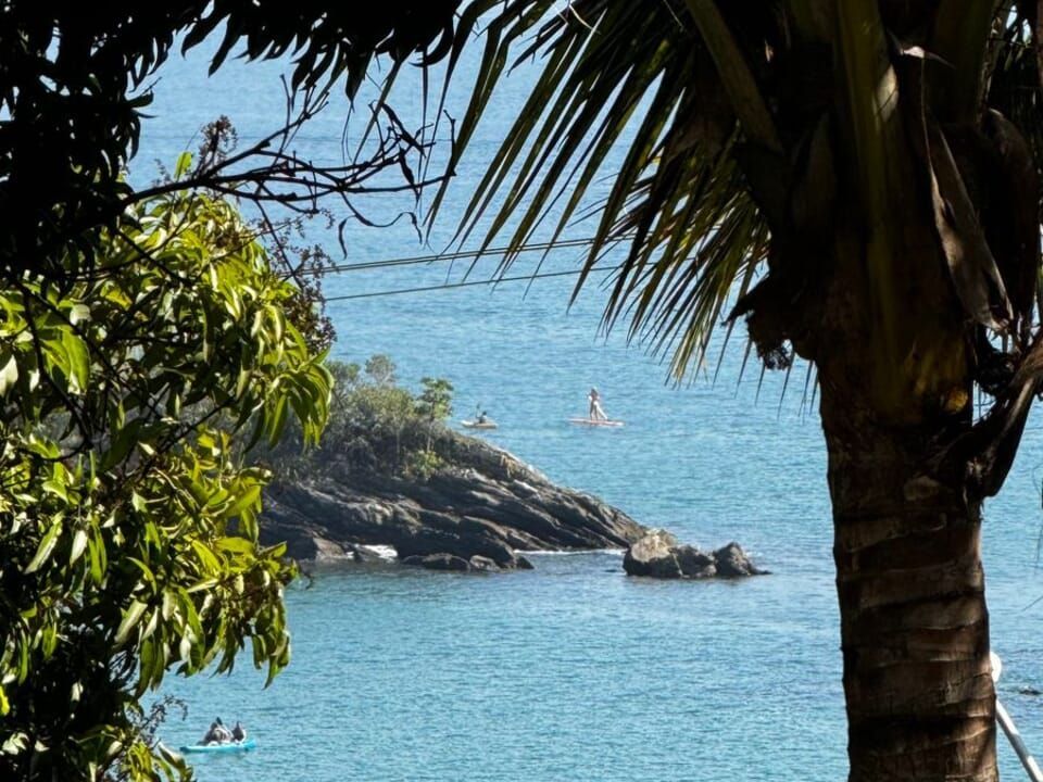 Uma vista para o mar azul e um promontório rochoso emoldurado por árvores, com pessoas praticando stand-up paddle e caiaque na água.