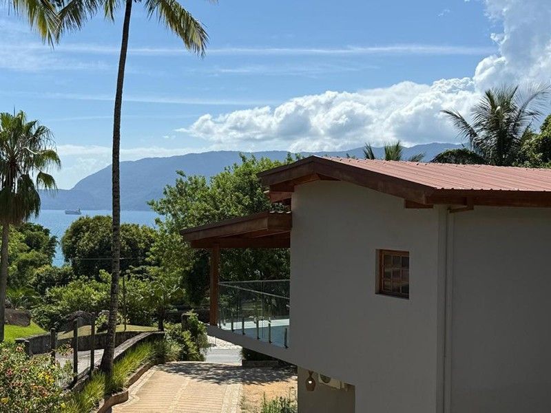 Uma casa moderna com um terraço com vista para uma costa tropical, árvores verdejantes e montanhas distantes sob um céu azul.