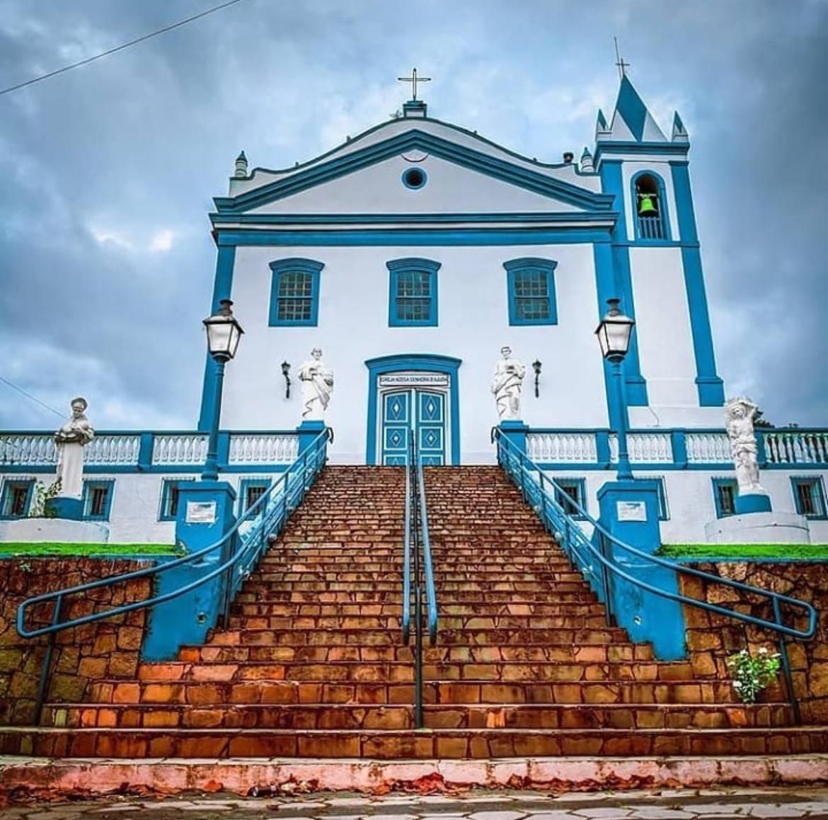 Uma igreja branca e brilhante com detalhes em azul vibrante, apresentando uma escadaria íngreme de pedra que leva à sua entrada sob um céu nublado.