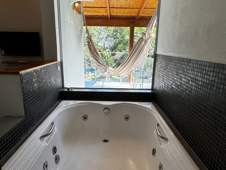 Uma banheira de hidromassagem branca encostada em paredes revestidas de azulejos pretos, em frente a uma grande janela com vista para um pátio com uma rede.