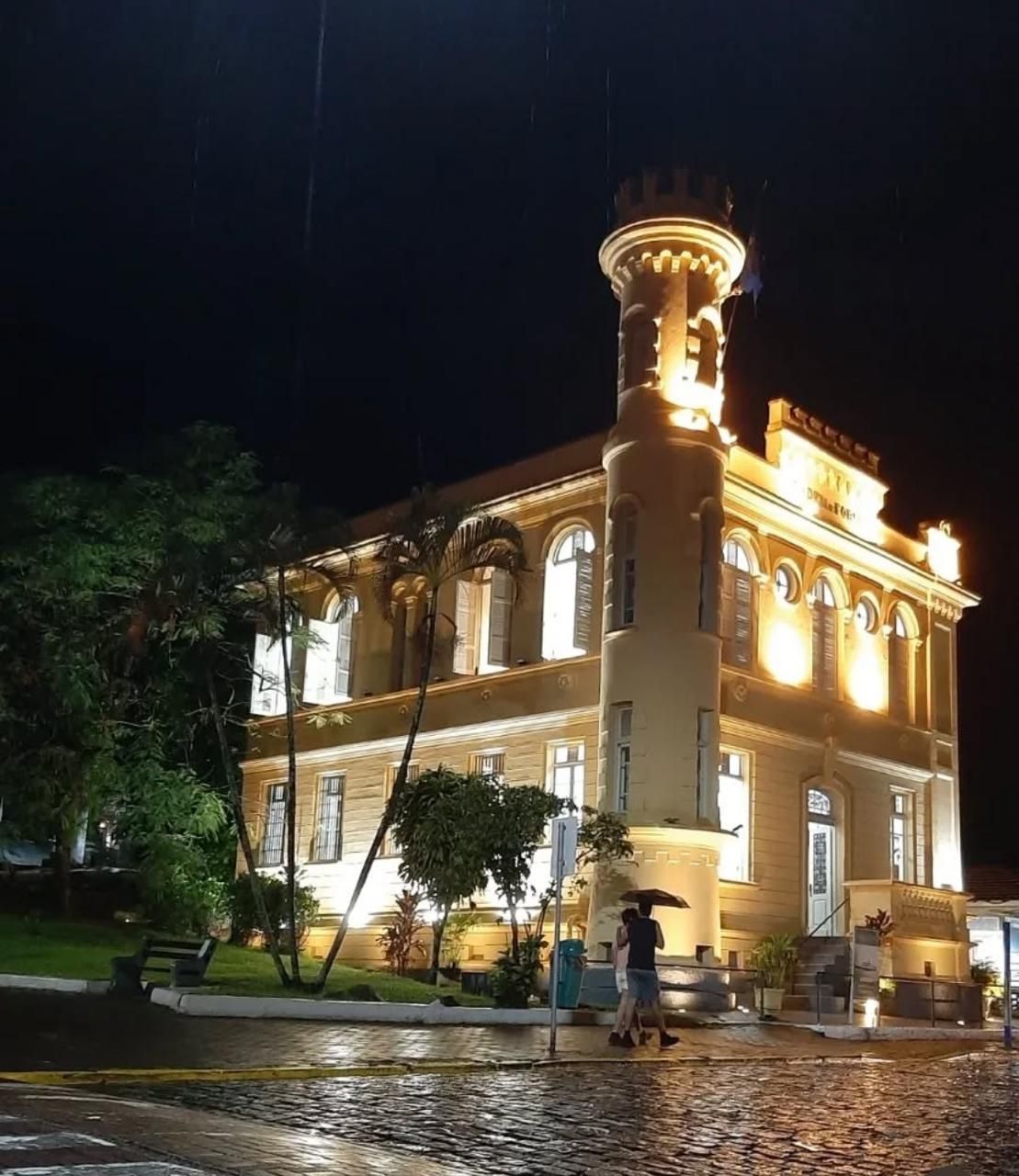 Vista noturna de um prédio histórico amarelo de dois andares com uma torre de esquina proeminente, iluminado contra um céu escuro.