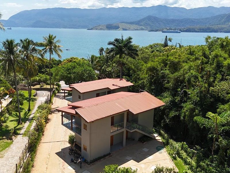 Vista aérea de um prédio de dois andares em tom bege com telhado de terracota, aninhado em meio a árvores verdejantes com vista para uma baía pitoresca.