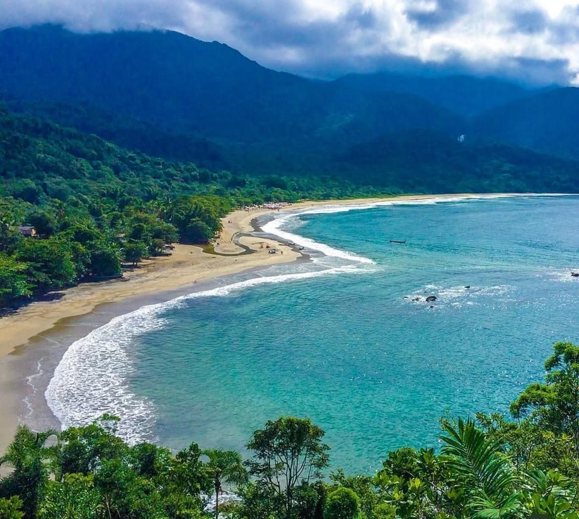 Uma praia tropical com água turquesa e ondas que encontram uma faixa de areia, tendo como pano de fundo montanhas verdejantes.