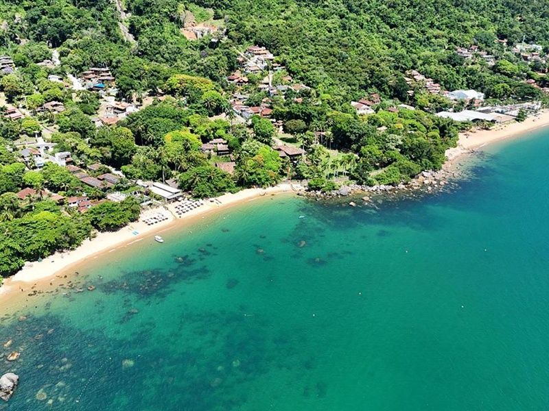 Vista aérea de uma praia de areia banhada por um mar azul-turquesa cristalino, emoldurada por densas árvores verdes e uma pequena cidade na encosta de uma colina.