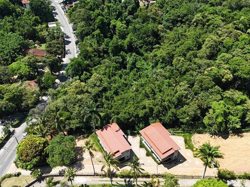 Vista aérea de duas casas com telhados vermelhos situadas na orla de uma densa floresta, ao lado de uma estrada sinuosa.