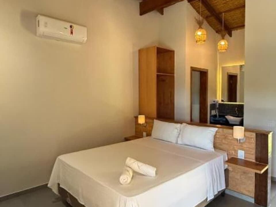 Um quarto de hotel moderno com uma cama queen-size, cabeceira de madeira clara, duas luminárias pendentes e um ar-condicionado branco na parede.