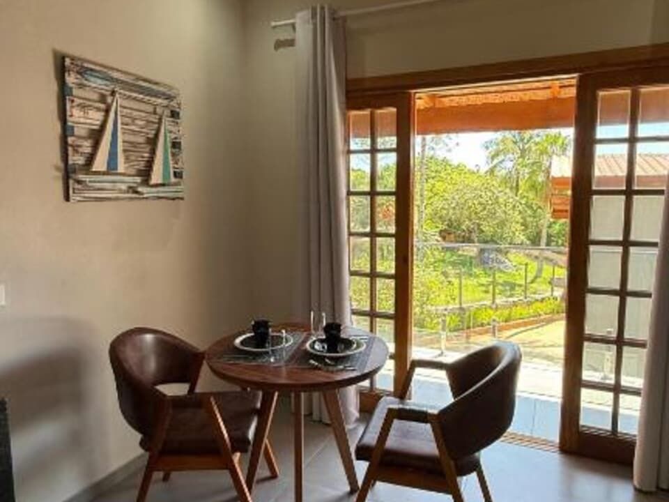 Uma pequena mesa de jantar de madeira com duas cadeiras de couro fica ao lado de uma porta de vidro deslizante que se abre para uma vista ensolarada do exterior.