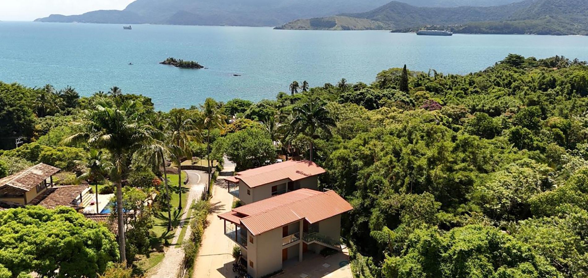 Duas casas com telhados de terracota aninhadas em uma paisagem costeira exuberante e verdejante, com vista para uma baía azul e montanhas ao longe.