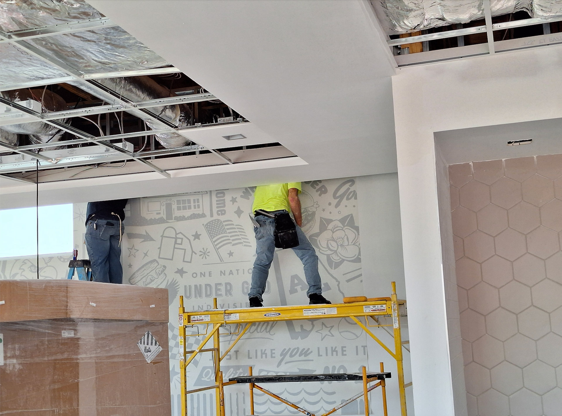 Workers installing ceiling panels in a building under construction. One stands on a scaffold, another is at the left.