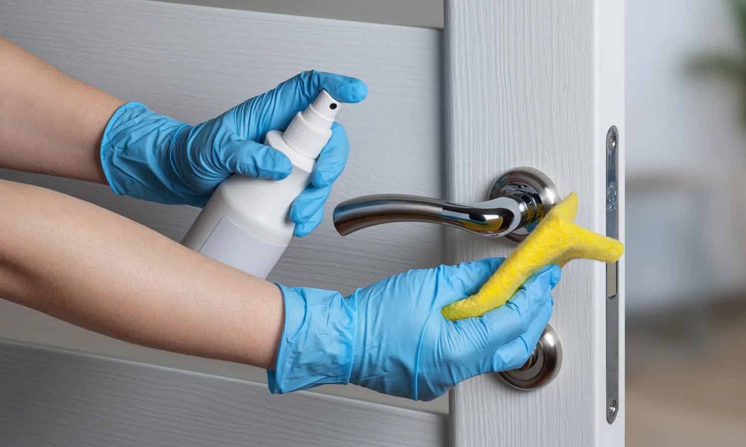 Une personne portant des gants bleus nettoie une poignée de porte avec un vaporisateur et une éponge.