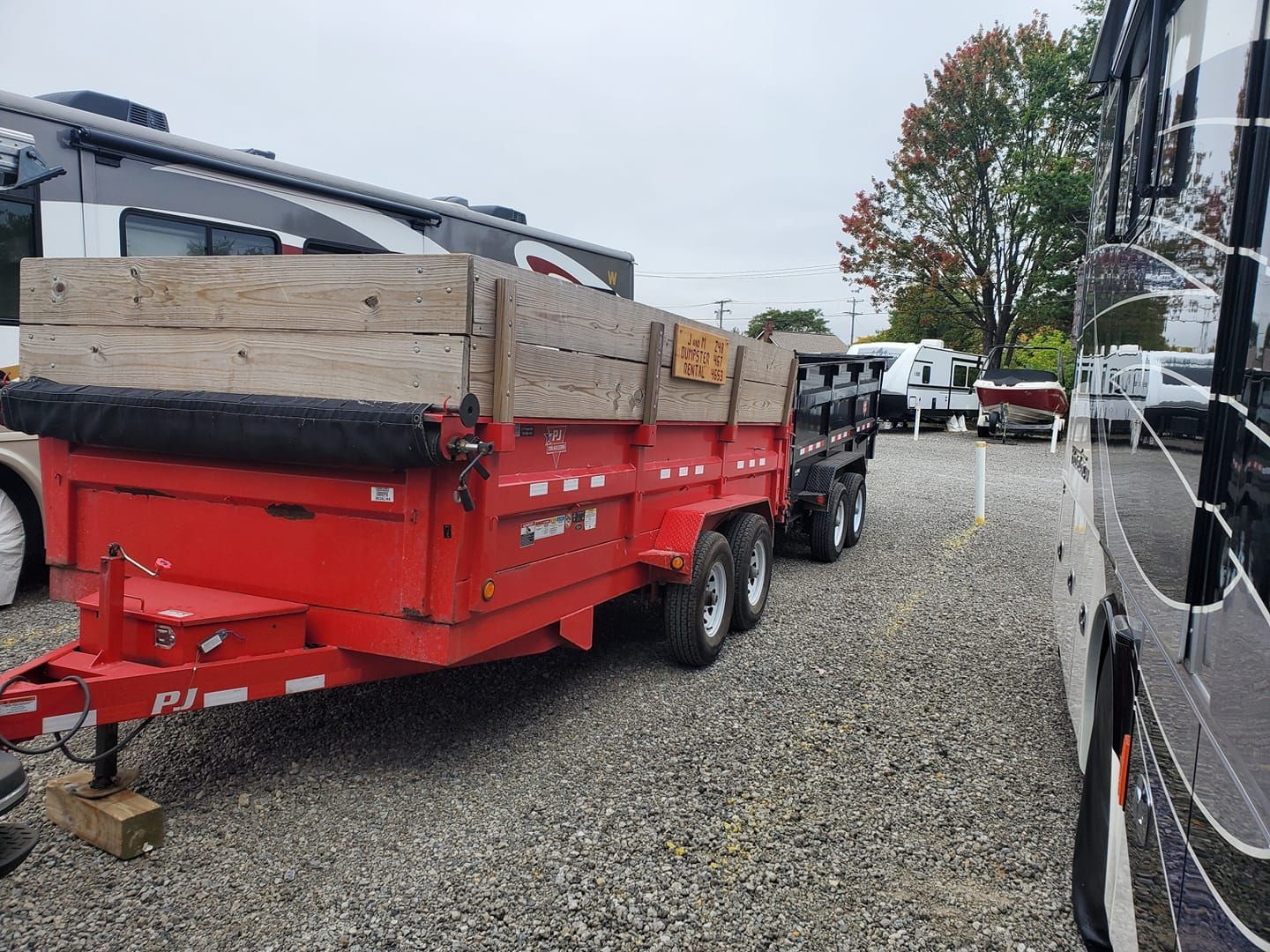 A red dump trailer is parked in a gravel lot next to a rv.