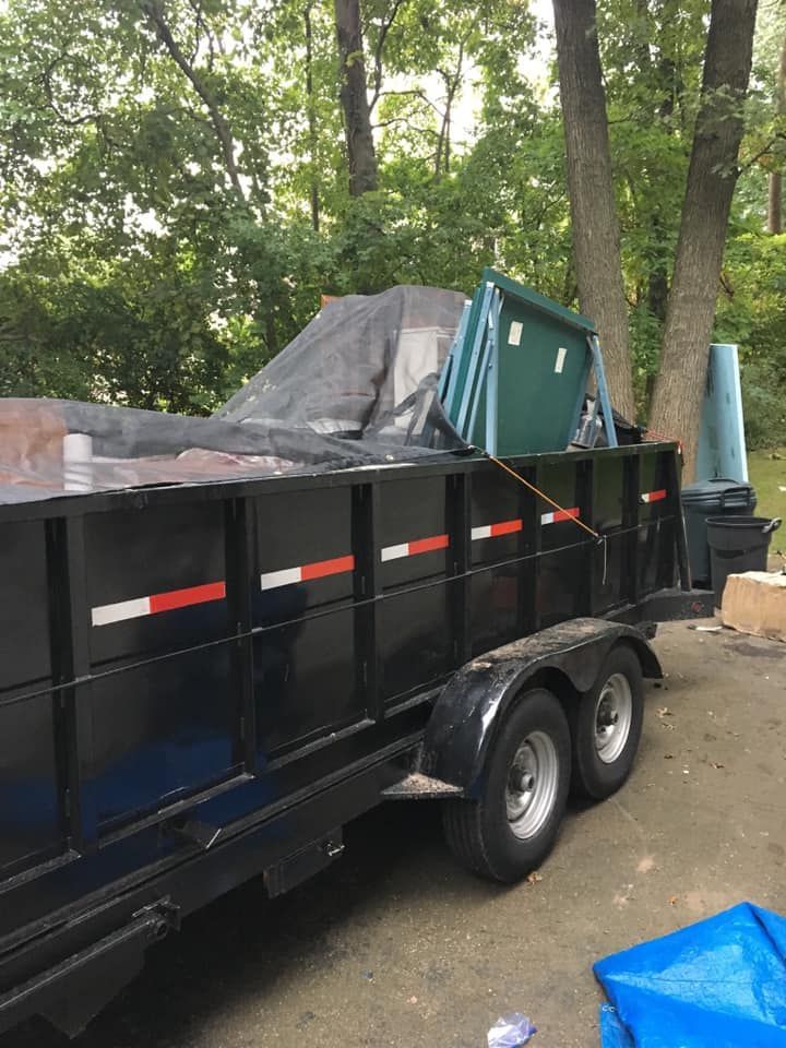 A dumpster trailer is parked in a driveway with trees in the background.