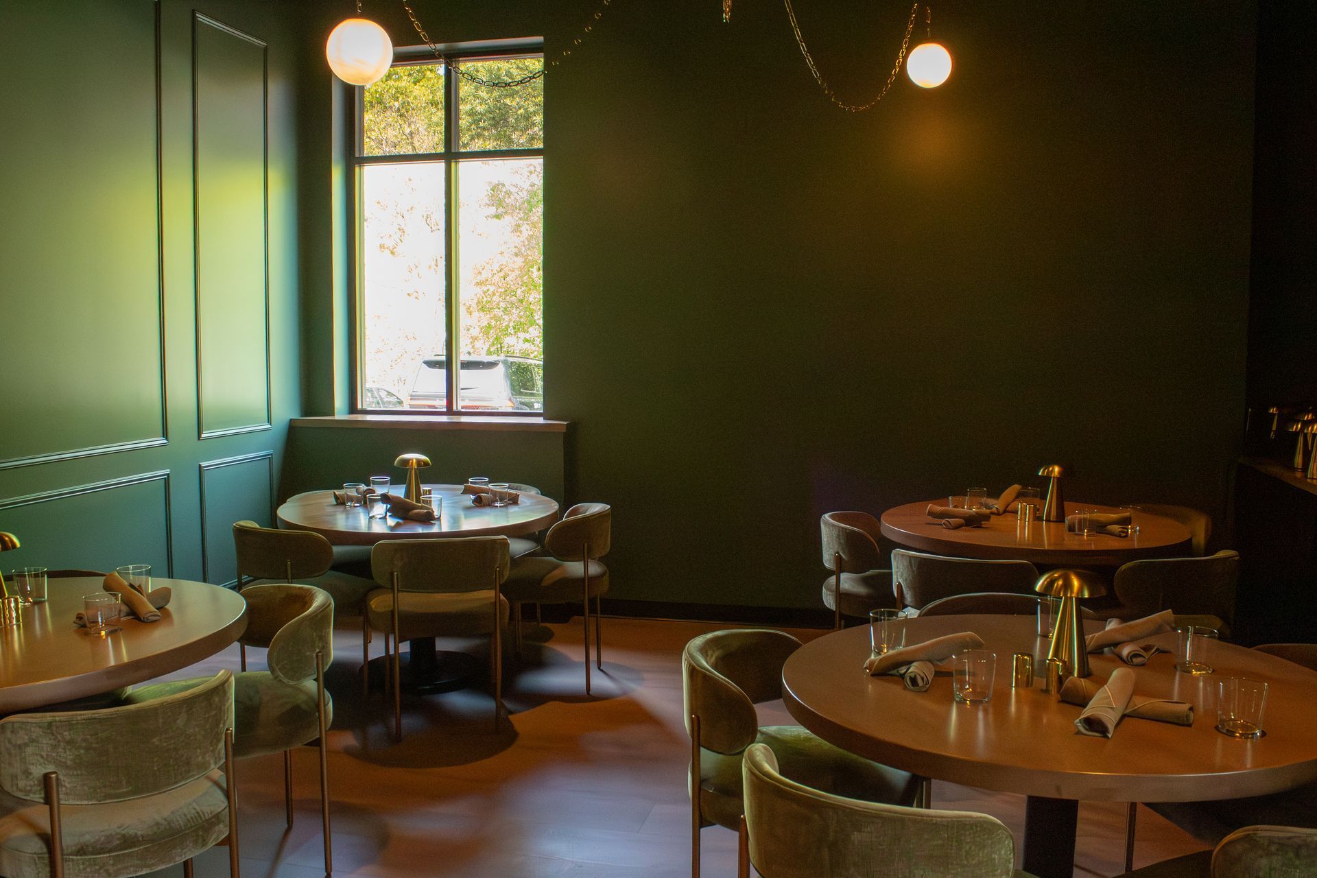 Dim restaurant dining room with round tables, dark walls, and a window with sunlight.
