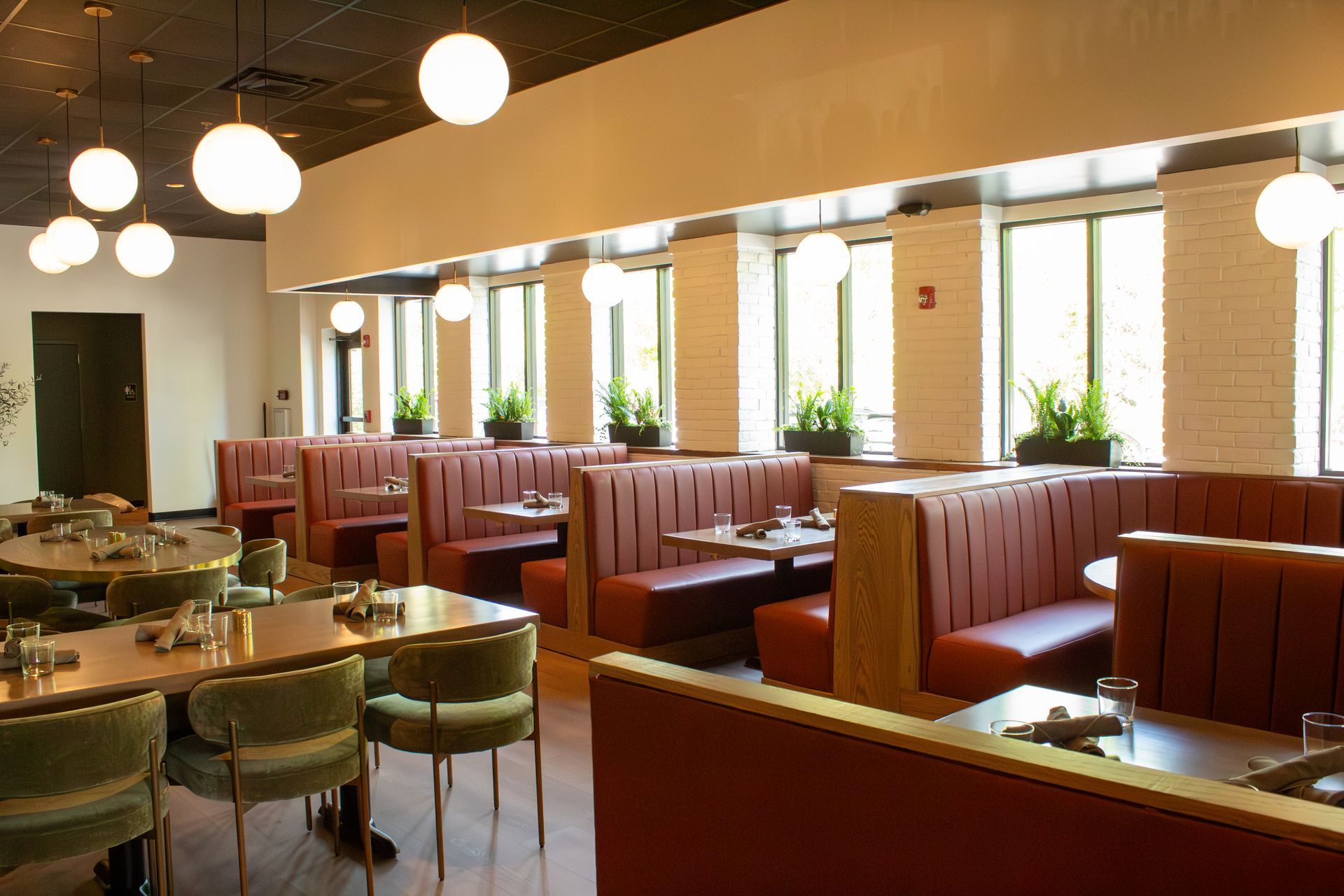 Bright restaurant interior with red booths, round tables, and hanging globe lights along tall windows.