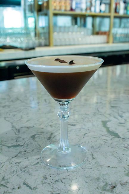 A dark cocktail in a textured glass, garnished with black olives on a bar counter.