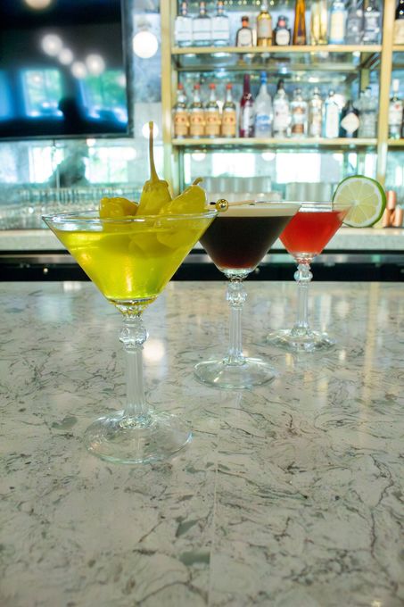 Three colorful cocktails on a marble bar with liquor bottles in the background