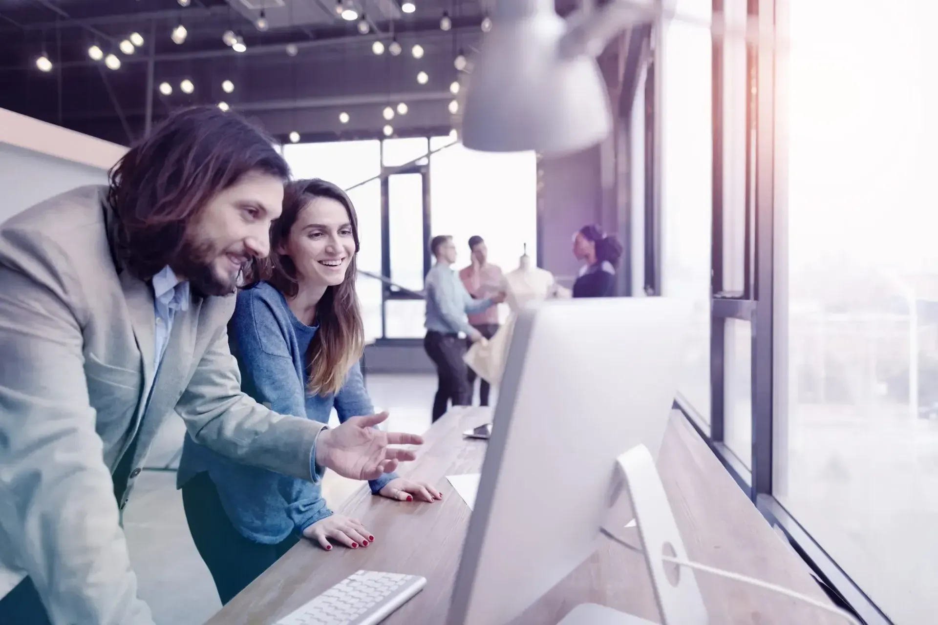 Two people looking at a computer screen in an office setting with other people in the background.