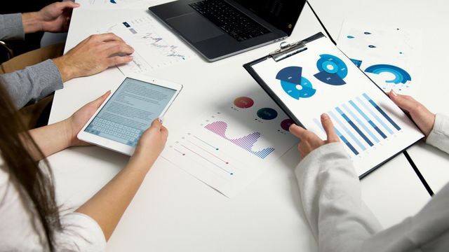 People around a white table, reviewing financial charts and data on a tablet and clipboard, with a laptop in the background.