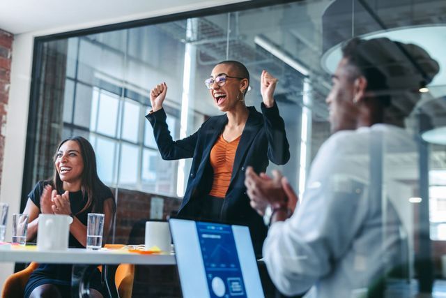 Businesspeople celebrating in a modern office; woman with arms raised, others clapping at a table with a laptop.