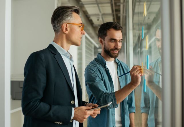 Two men at a glass wall with sticky notes, one writing with a pen, other holding a tablet.