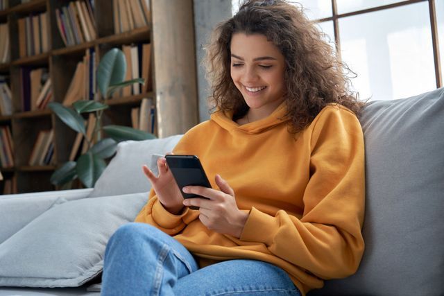Woman with curly hair smiles while using a smartphone, seated on a couch in front of a bookshelf. She is wearing a yellow hoodie and jeans.