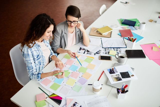 Two women at a table with sticky notes, documents, and a laptop, collaborating on a project.