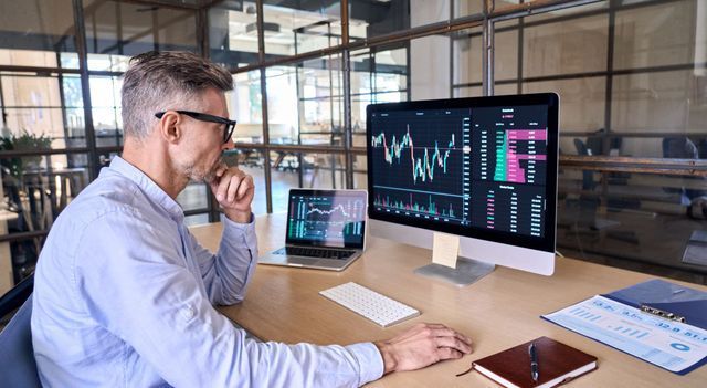 Man analyzing stock charts on multiple screens in an office setting, with a serious expression.
