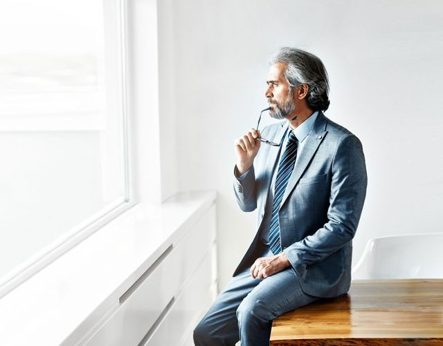 Man in blue suit, sits on table, looks out window, holds glasses.