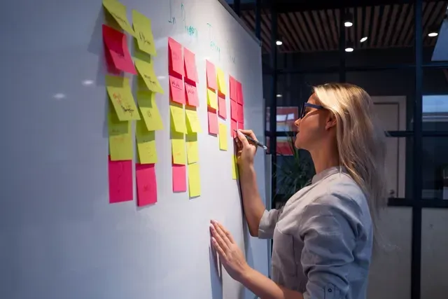 Woman writing on pink sticky note on a whiteboard covered in yellow and pink sticky notes.