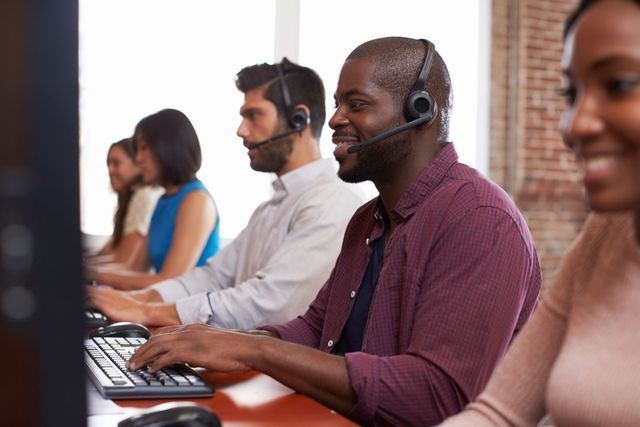 People in a call center, wearing headsets, typing at computers, and smiling.