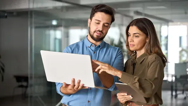 Man and woman looking at a laptop together, pointing at the screen in an office setting.