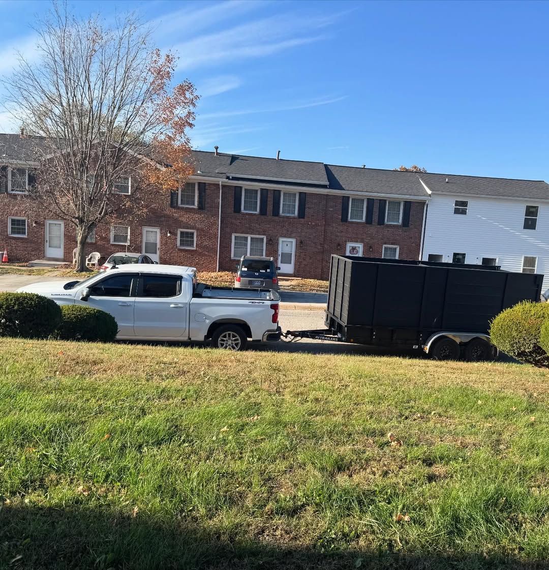 White truck towing a black dumpster trailer parked on a grassy lawn in front of townhouses.