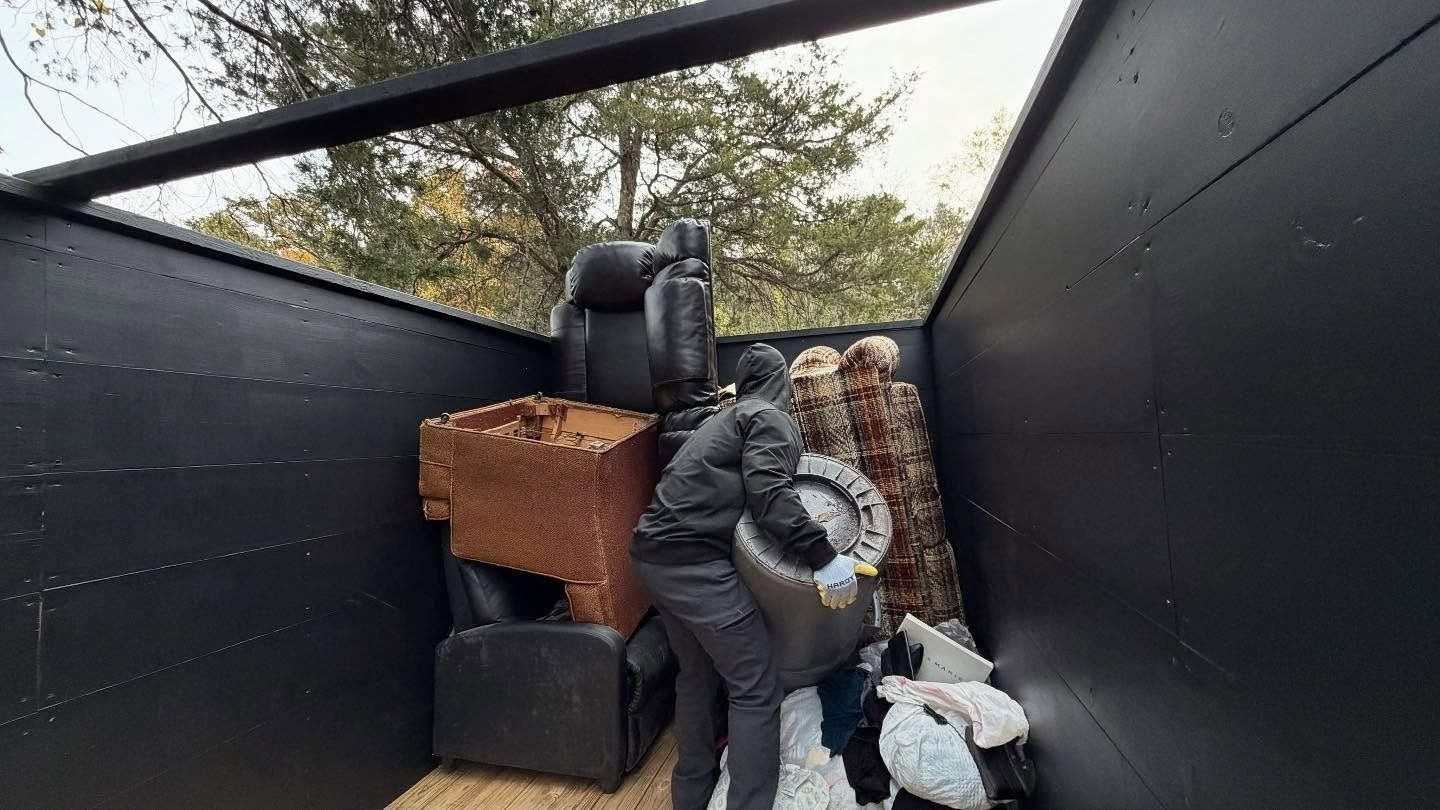 Person loading trash into a black trailer; contains furniture and debris. Outdoors setting.