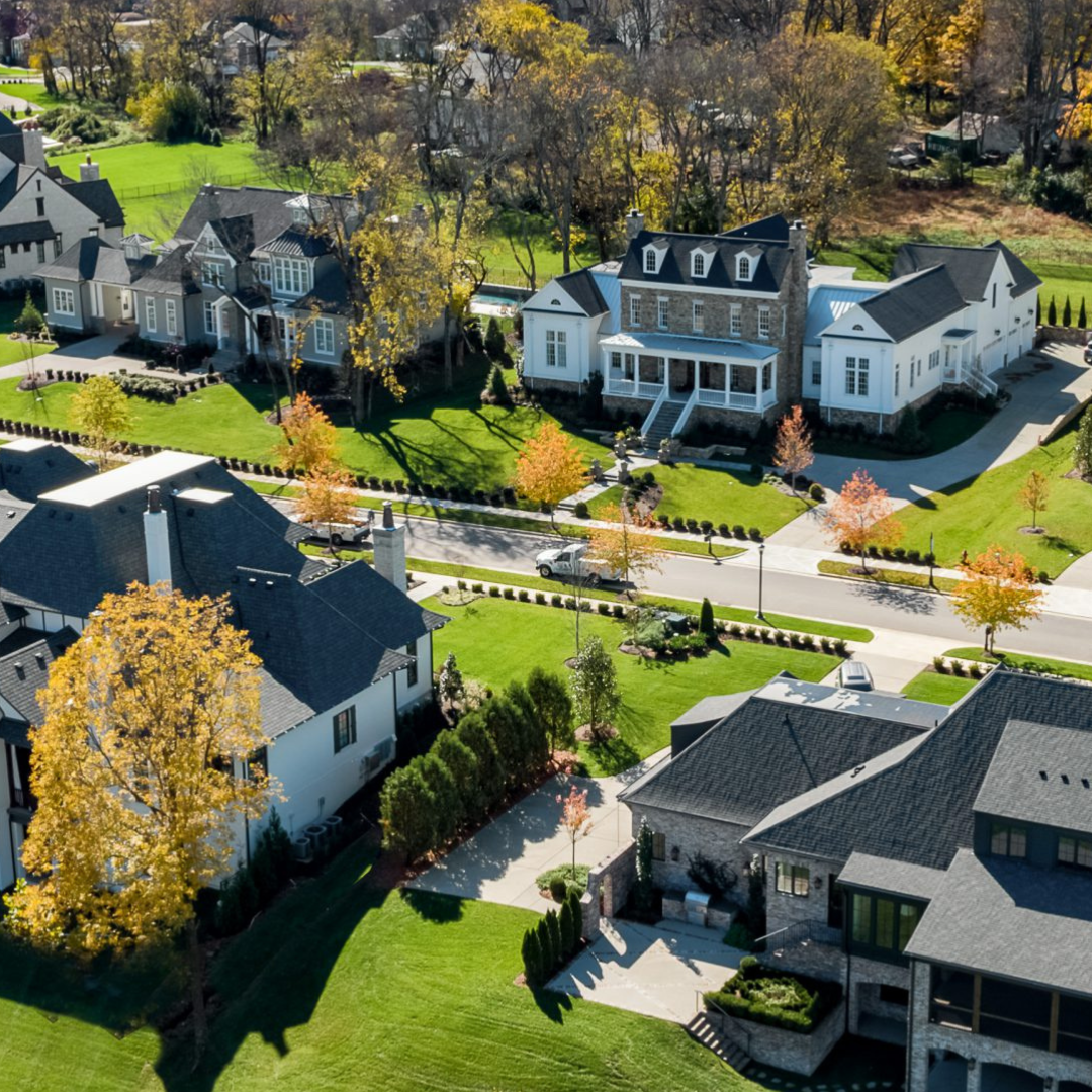 Aerial view of suburban homes with manicured lawns, autumn trees, and a winding street lined with decorative hedges.