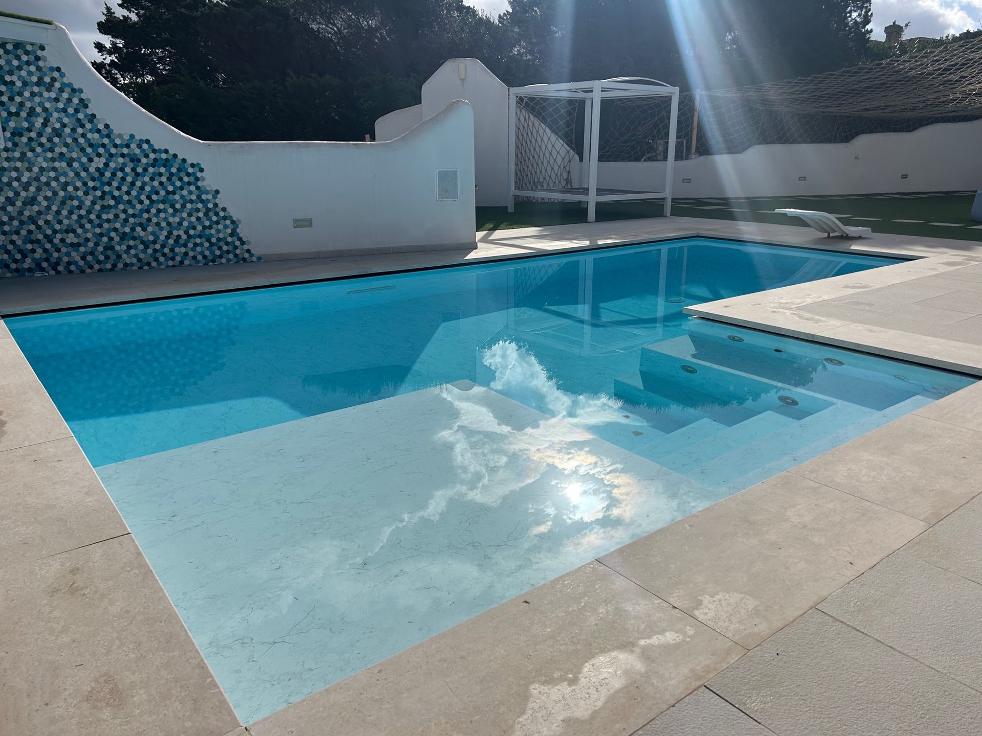 Una piscina rettangolare di un blu brillante con scalini integrati, circondata da un patio di colore chiaro e pareti bianche.