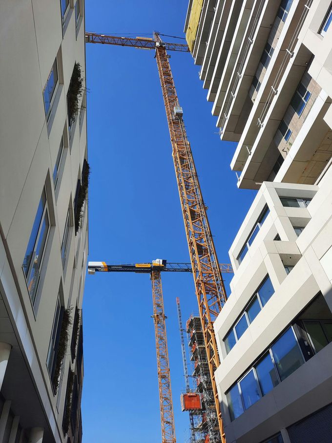 Vista dal basso di due alte gru edili che si ergono tra edifici moderni, sullo sfondo di un cielo azzurro e limpido.