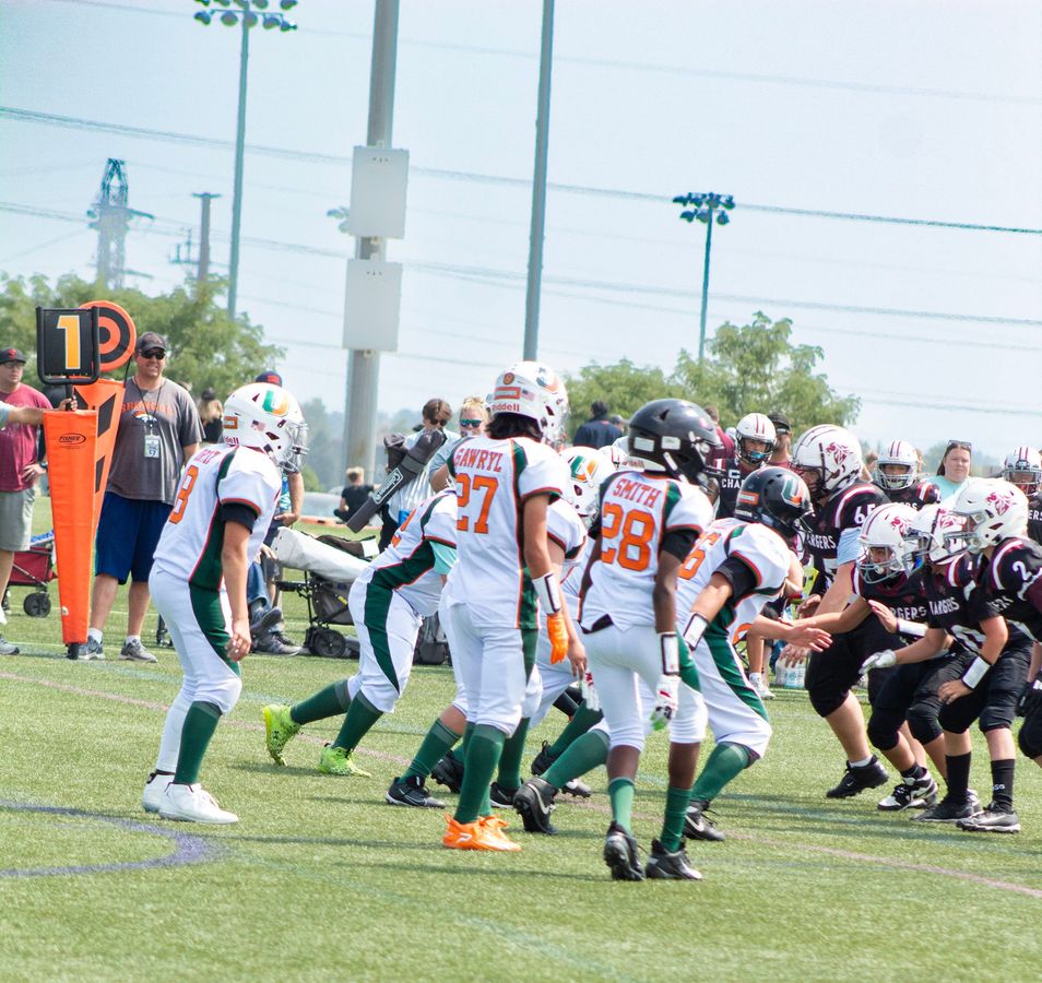 Football players in action on a grassy field; one runs with the ball, pursued by two opponents.