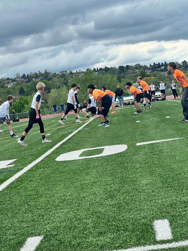 Flag football game at night, a player in red jersey runs with the ball as others pursue on a green field.