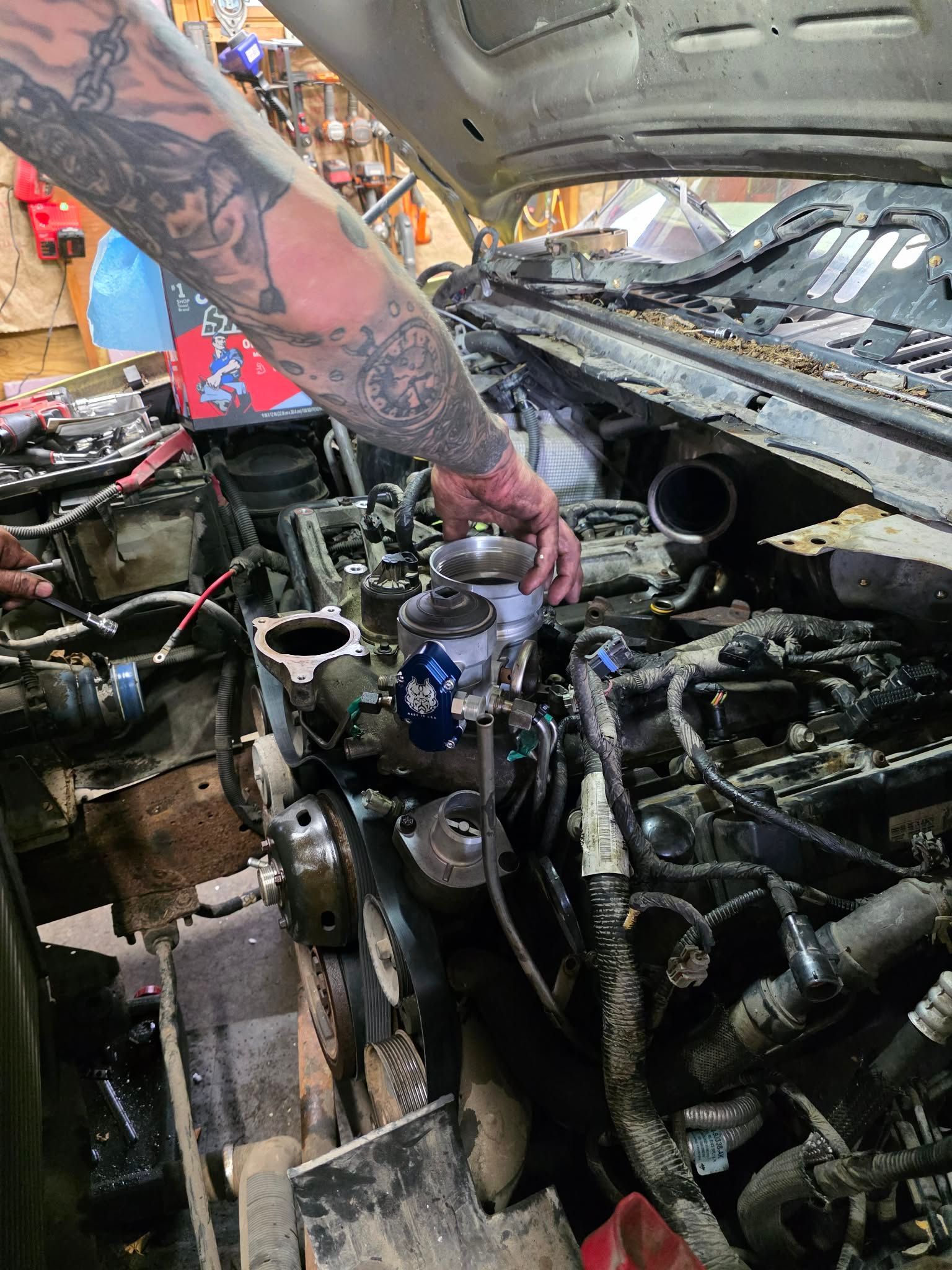 Mechanic working on a car engine, arm covered in tattoos, visible engine parts and tools, indoor setting. | Seth's Auto Care